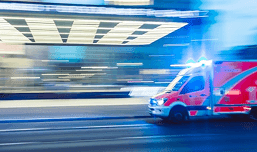 A blurred city street shows a red ambulance entering the frame from the right.