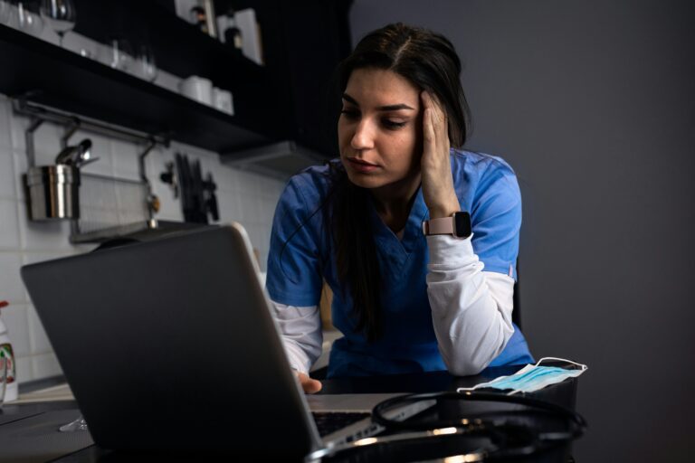 A frustrated healthcare provider rubs their temple while looking at a laptop in the hospital.