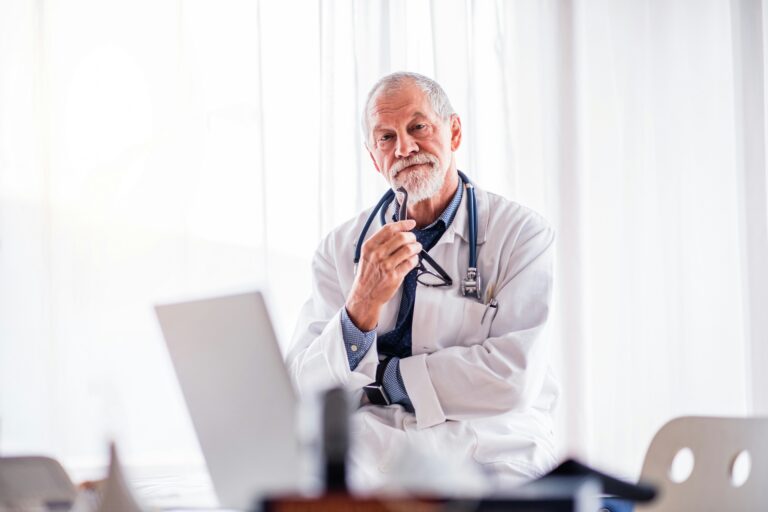A white-haired doctor looks pensively at the camera with his reading glasses against his chin.