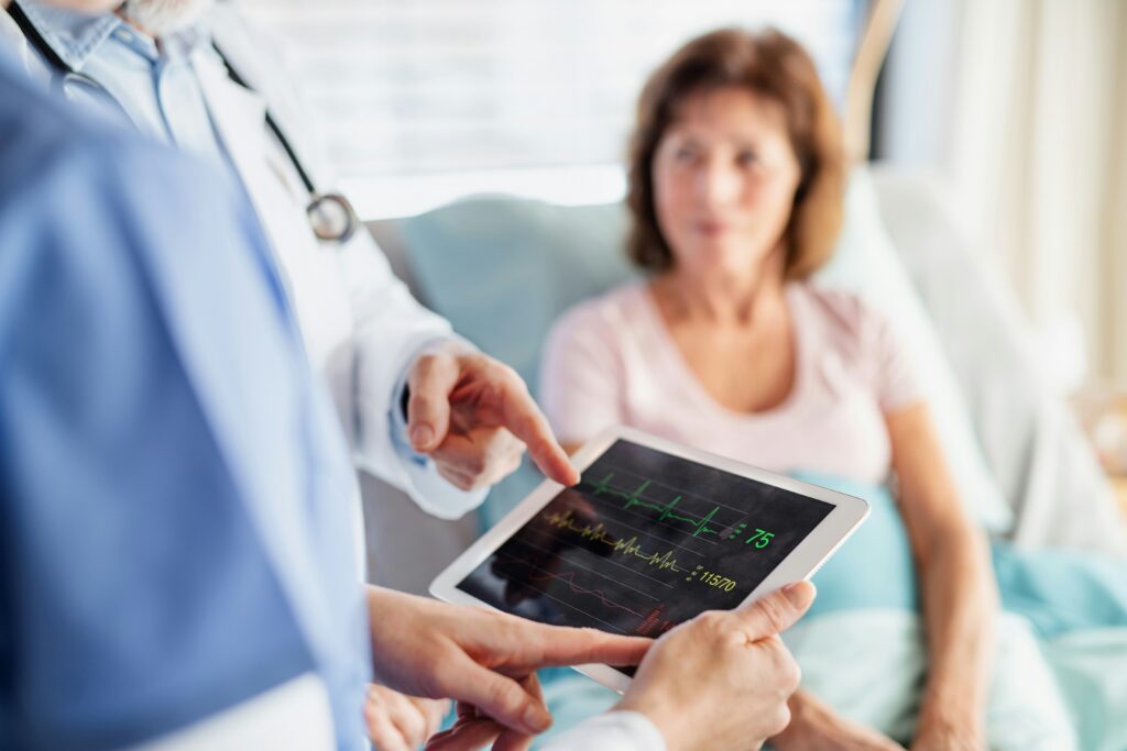 In the foreground, healthcare providers' hands point to a digital pad with health data on it. In the background, a hospital patient lays in a bed.