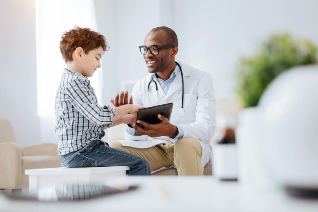 A physician laughs with a young patient who pokes the physician's tablet.