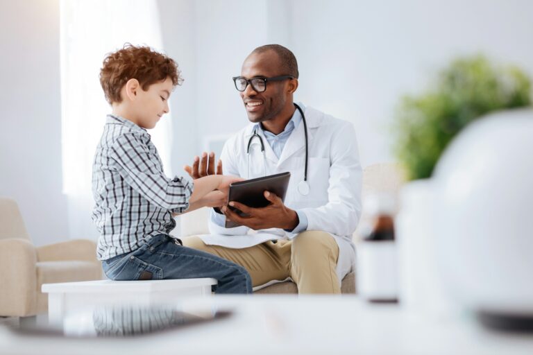 A physician laughs with a young patient who pokes the physician's tablet.