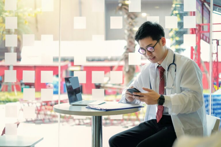 A young doctor smiles as he checks his phone with a laptop and clipboard on a table in front of him before a large bright window.