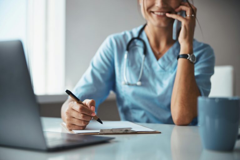 A doctor is smiling while on the phone with a pen in their hand and a clipboard and a laptop in front of her.